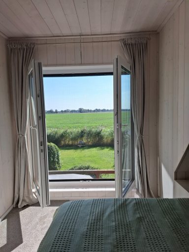 Blick aus einem Zimmer mit offenem Fenster auf eine grüne Wiese und klarem Himmel.