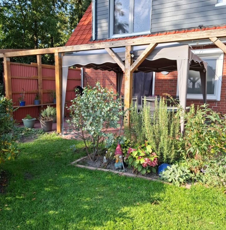 Terrasse mit Tisch und Stühlen unter Pavillon im Sommerlicht Holzgartenlaube mit Pflanzen und bunten Blumentöpfen im grünen Garten.