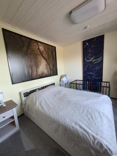 Helles Schlafzimmer mit einem Bett, Nachttisch, Gitterbett und Kunstwerken an der Wand.