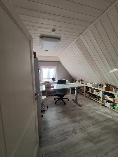 Ein heller, schrägwandiger Raum mit Schreibtisch, Stuhl und Regalen voller Gegenstände.