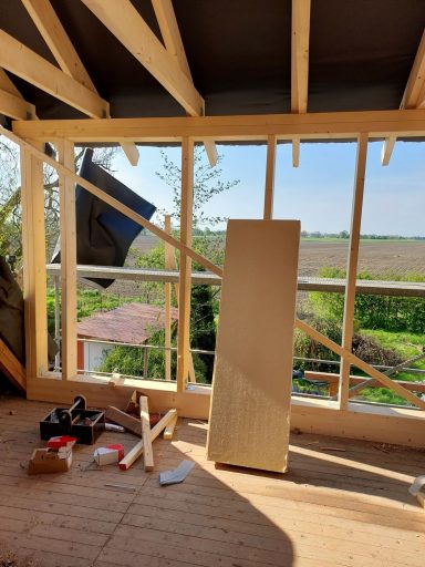 Bau der Dachgaube Blick auf einen unvollendeten Raum mit Holzrahmen und Fenstern, Baustellenmaterialien im Vordergrund.