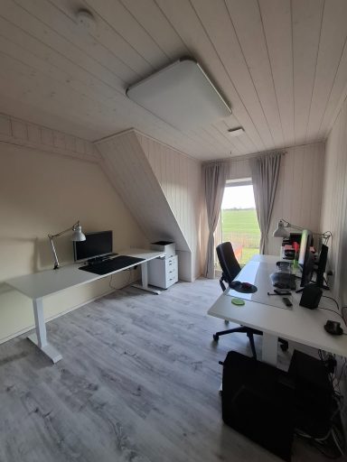 Helles Arbeitszimmer mit Schreibtisch, Monitoren und Fenster mit Blick ins Freie.