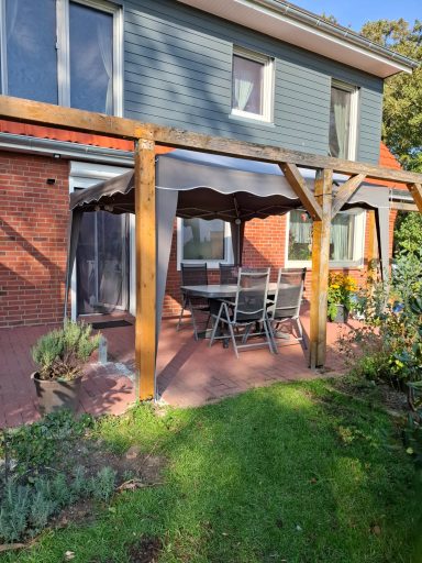 Terrassenbereich Überdachte Terrasse mit Tisch und Stühlen, umgeben von Pflanzen und Rasen.